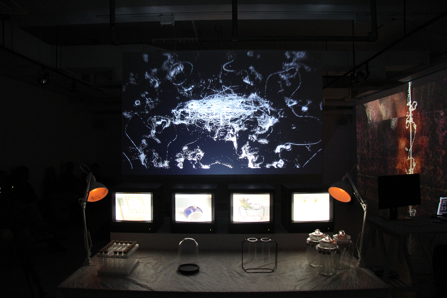 Image de l'exposition. Dans une ambiance sombre, quatre écrans de télévision sont alignés et projettent une lumière blanche. De part et d'autre, deux lumières de bureau orange encadrent les écrans. Au-dessus des télévisions, un plus grand écran affiche des formes grisâtres aux allures futuristiques.