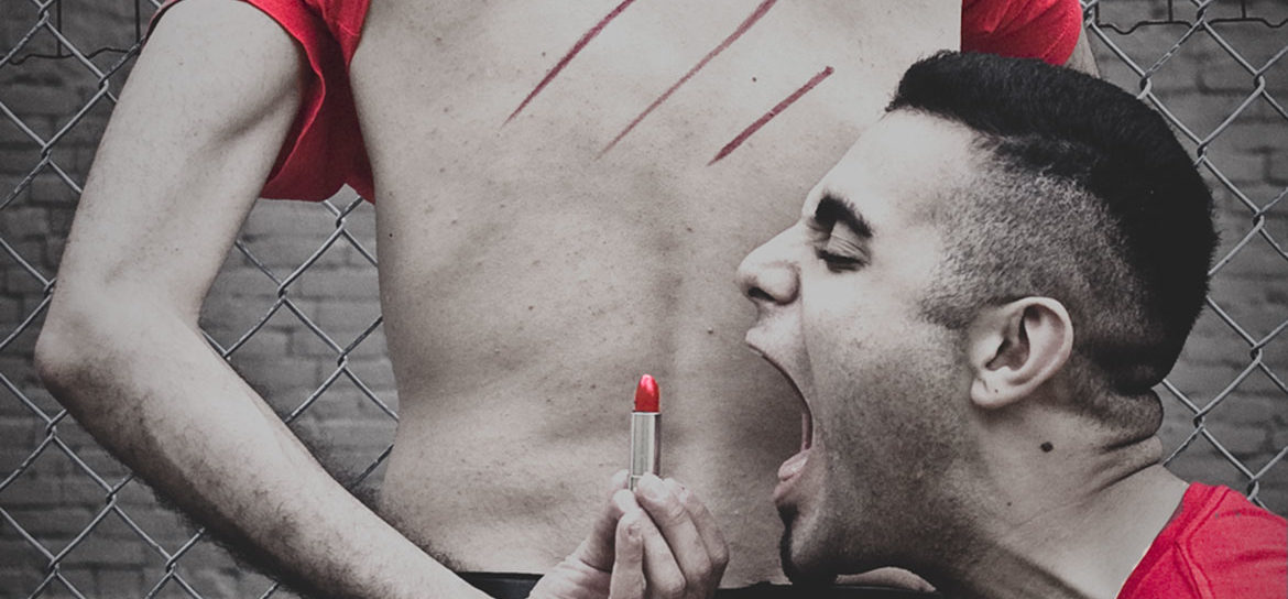 Person with back turned towards viewer. Red t-short pulled over their head. Three diagonal red lipstick lines drawn on thier back. A second person with their mouth wide open, about to eat lipstick, held by first person.