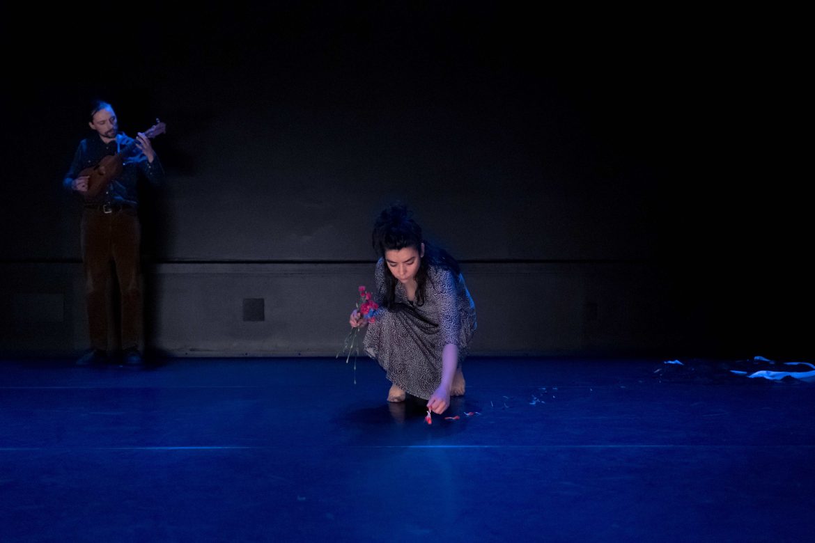 Image du spectacle Je ne vais pas inonder la mer.
Au centre, Sonia Bustos est accroupie et porte une robe à fleurs grise. Elle répand des pétales de fleur rose sur le sol. En arrière-plan à gauche, un musicien joue d'un instrument à corde ressemblant à une petite guitare.